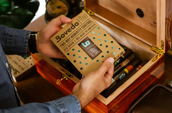 Hands holding and placing a 69% RH Size 320 Boveda into a medium wood humidor for storing cigars.