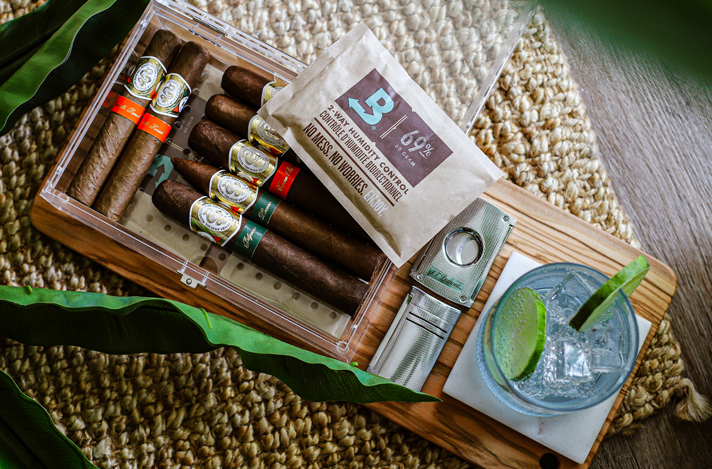 Small acrylic humidor for storing cigars with a 69% RH Size 60 Boveda, a cigar cutter, lighter and beverage.