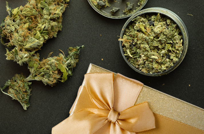 Cannabis buds and ground cannabis in a grinder on a table top with a present with golden wrapping and bow.