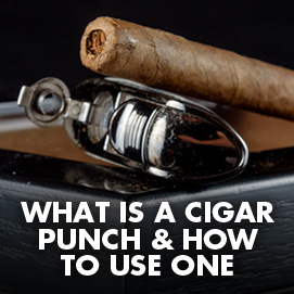HEADLINE: What is a Cigar punch & how to use one. IMAGE: Single cigar with a punch in the cap along with a cigar punch device sitting on a humidor box.