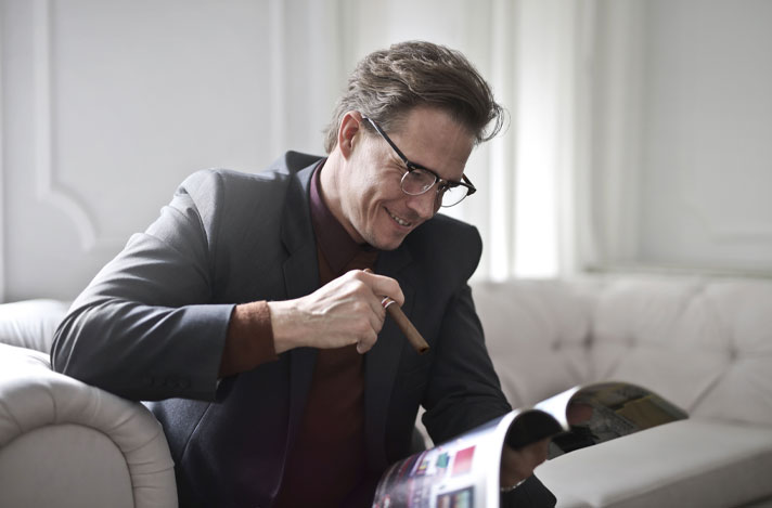 A man sitting on a couch reading a magazine while smoking a cigar